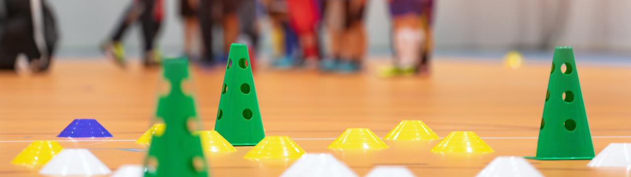 Bunte Hütchen auf Hallenboden. Kinder und Trainer verschwommen im Hintergrund.