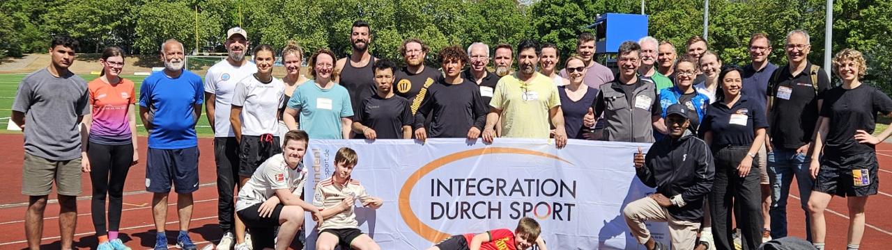 Das Foto zeigt alle Teilnehmenden des Netzwerktreffens auf der Bezirkssportanlage Mainz Mombach. Die Gruppe hält ein Banner mit dem Logo von "Integration durch Sport".