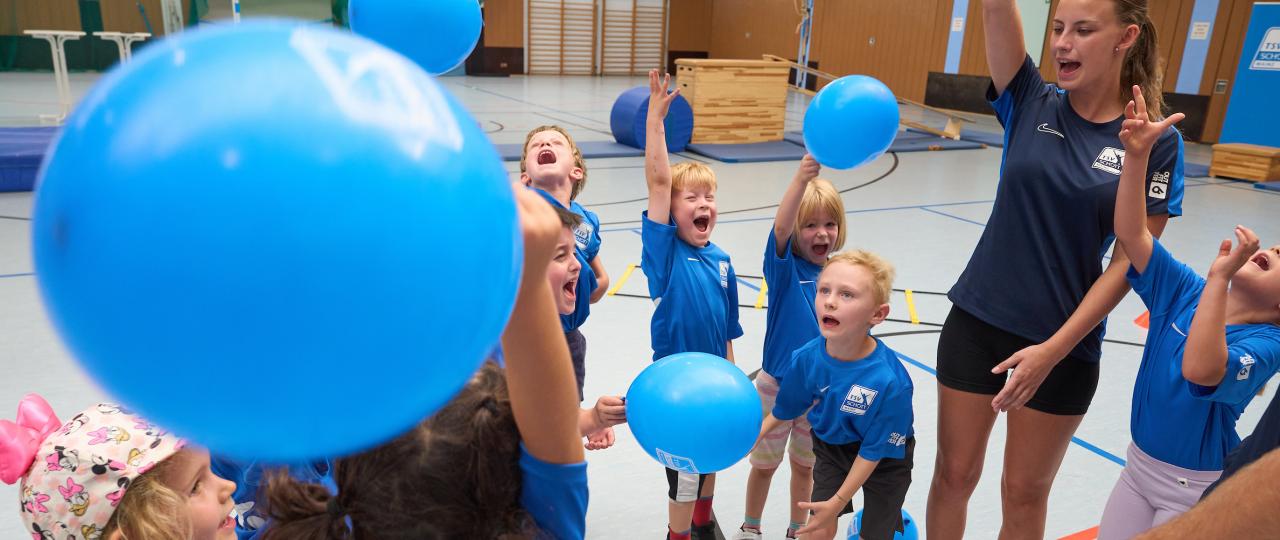 Kinder und junge Frau in Turnhalle mit blauen Luftballons