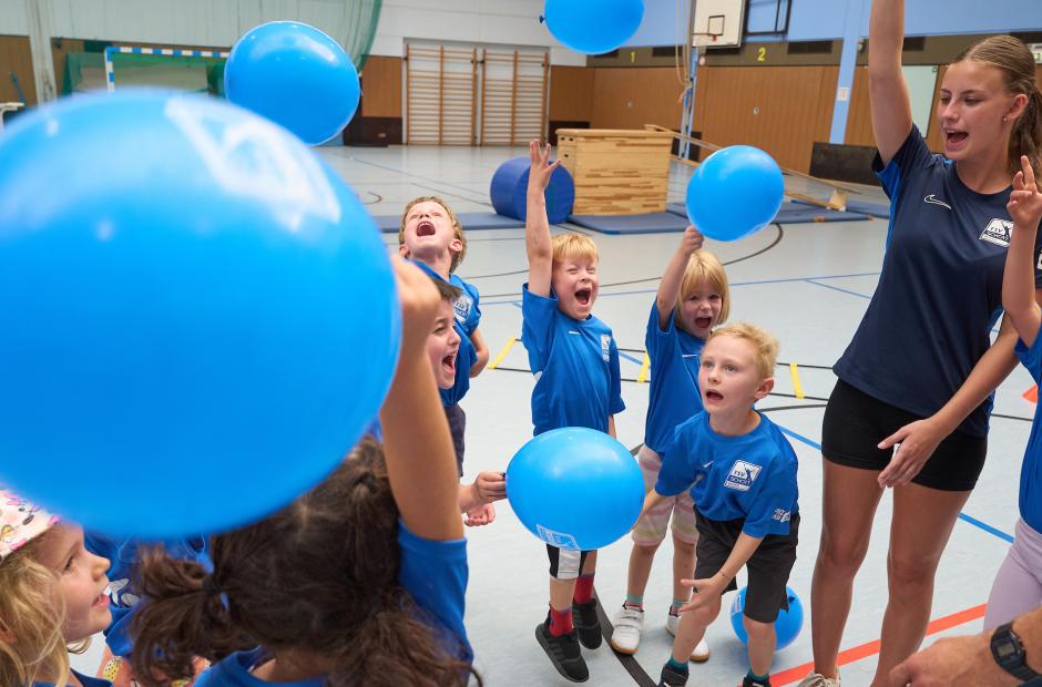 Kinder und junge Frau in Turnhalle mit blauen Luftballons