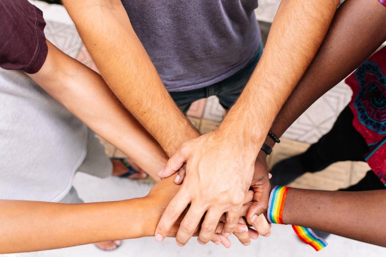 Mehrere unterschiedliche Hände übereinander gefaltet, eine Hand trägt dabei ein Symbol der LGBTQ-Bewegung. 