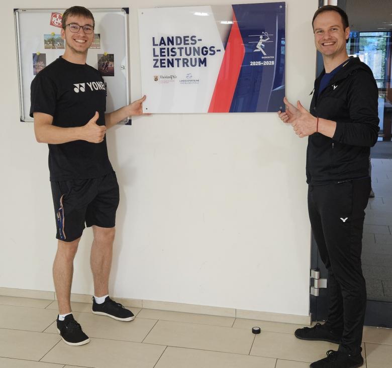 Zwei Männer vor Schild in Sporthalle