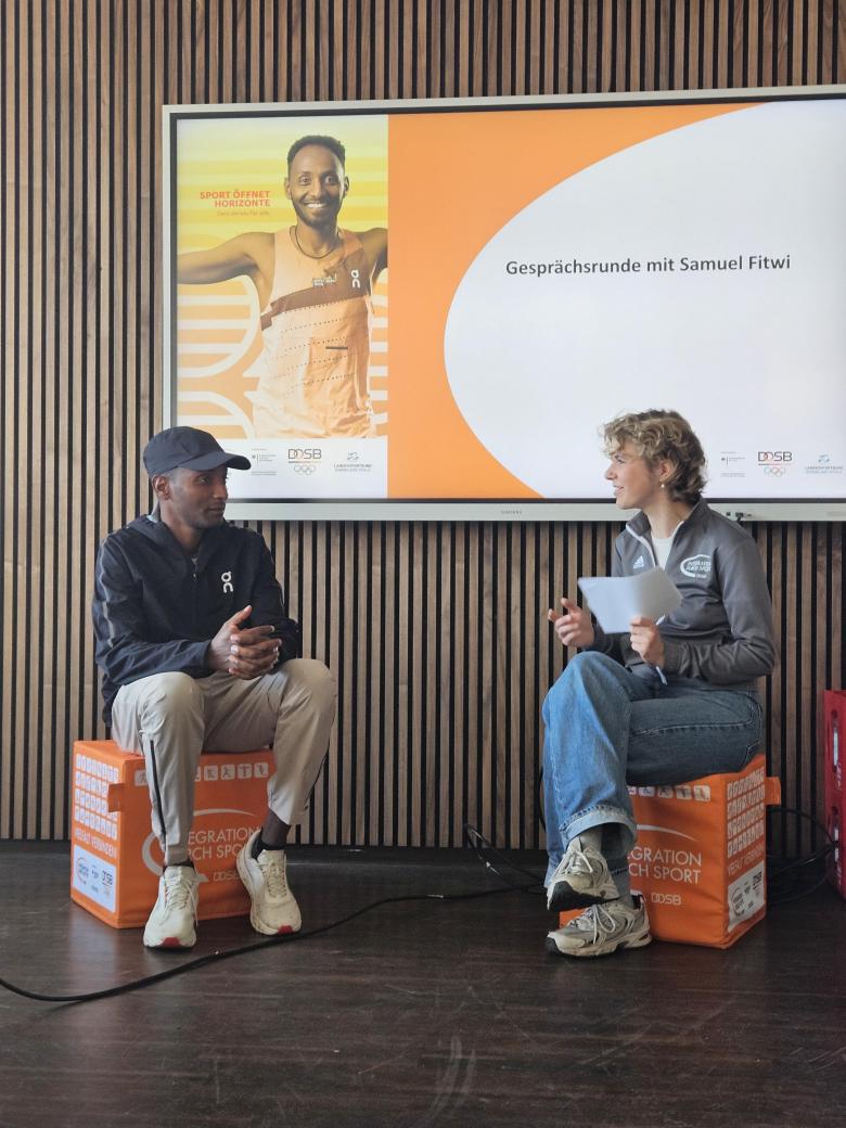 Die Programmreferentin Larissa Rohr und Samuel Fitwi sitzen auf zwei "Integration durch Sport"-gebrandeten Sitzkissen uns unterhalten sich.