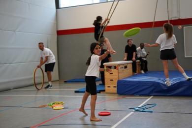 Mädchen in der Turnhalle. Im Vordergrund ein Mädchen, das eine Frisbeescheibe wirft. Im Hintergrund spielen sie auf Matten und an Seilen.