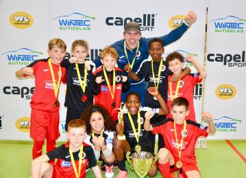 Das Foto zeigt ein Team junger Sportler*innen zusammen mit einem Trainer. Alle Personen tragen Medaillen und ein Kind hält einen Pokal in der Hand. Die gezeigten Personen jubeln über ihren Erfolg.