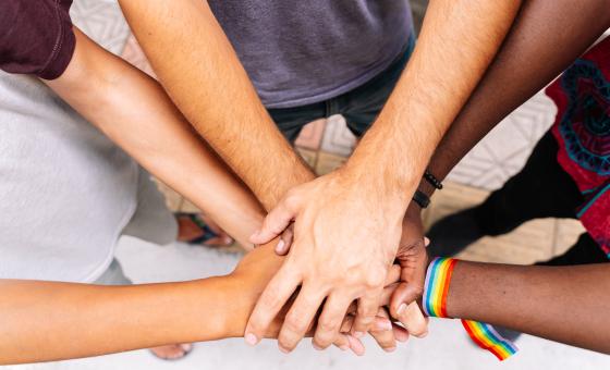 Mehrere unterschiedliche Hände übereinander gefaltet, eine Hand trägt dabei ein Symbol der LGBTQ-Bewegung. 