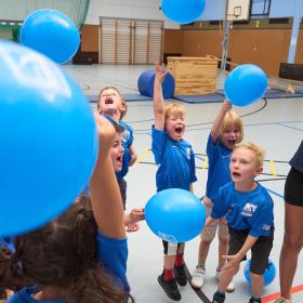 Kinder und junge Frau in Turnhalle mit blauen Luftballons